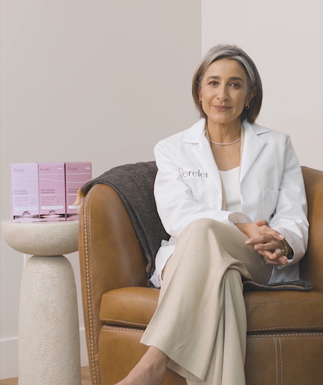 Woman in a white coat sitting on a brown chair with pink boxes in the background
