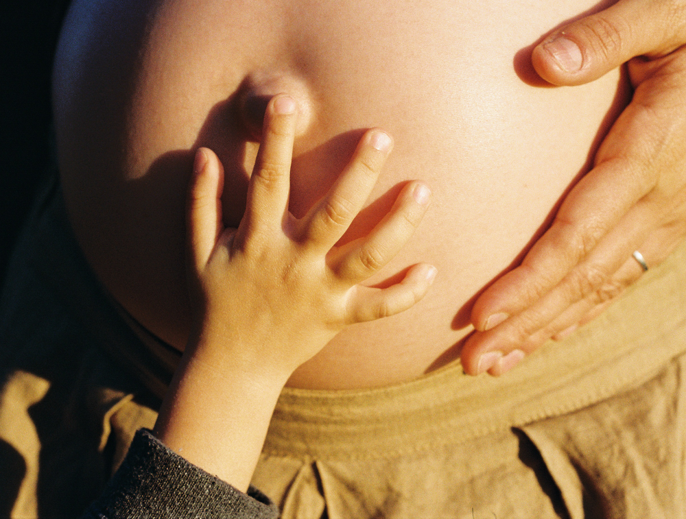 A child’s hand rests on a pregnant belly beside an adult’s hand.