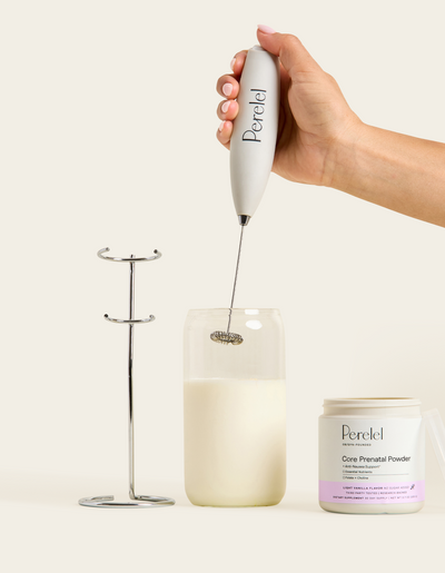 Hand holding a Perelel frother over a glass of creamy liquid, with a metal frother stand and an open jar of Perelel Core Prenatal Powder in light vanilla flavor beside it.